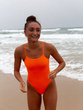 JAYD Swimwear features the Neon Orange Zimmy one piece from the front. This athletic one piece is a cheeky cut, it is being worn for leisure at the beach